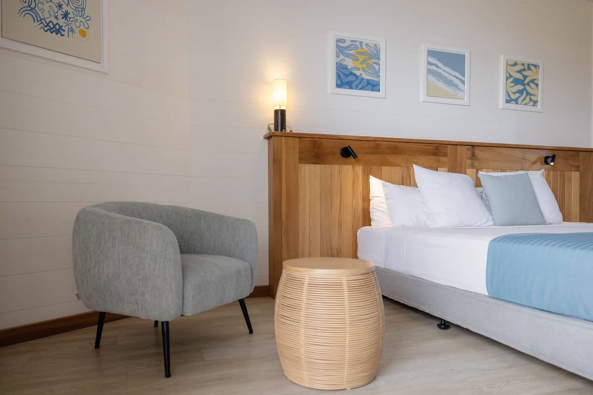 Un dormitorio con una cama, un sillón gris, un taburete de madera redondo y decoración en la pared.