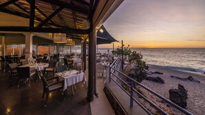 Un área de comedor al aire libre con mesas y sillas, con vista a una playa y al océano.