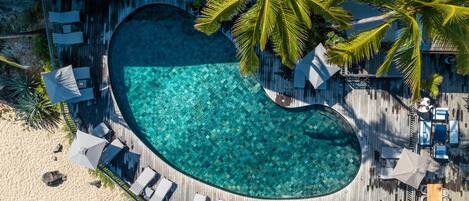 Vista aérea de un resort con piscina en forma de corazón, sillas de descanso y palmeras.