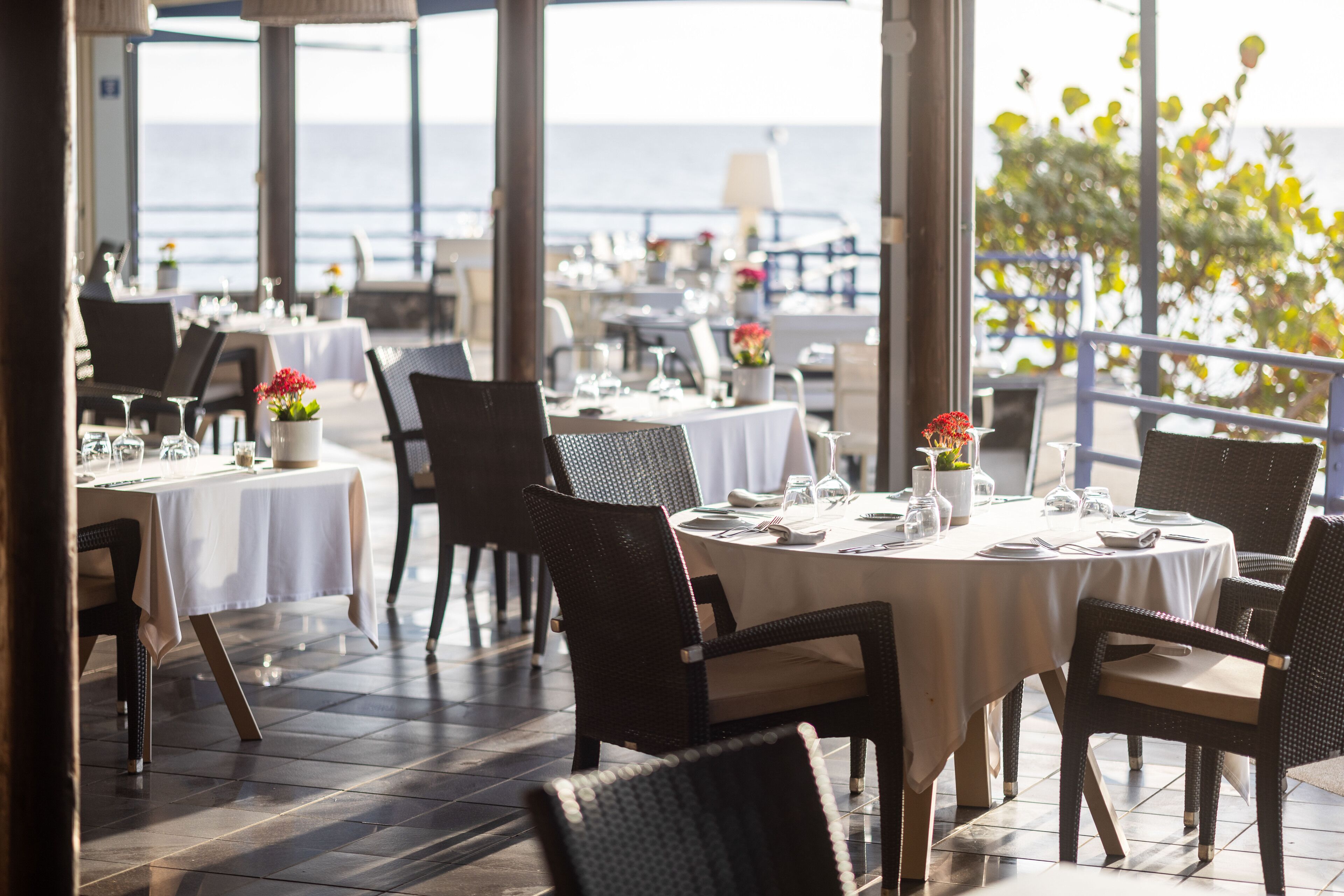 A restaurant with round tables and wicker chairs, set against a view of the sea.