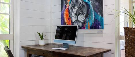 A wooden desk with a computer and a potted plant, a framed painting of a lion, and a wicker chair.