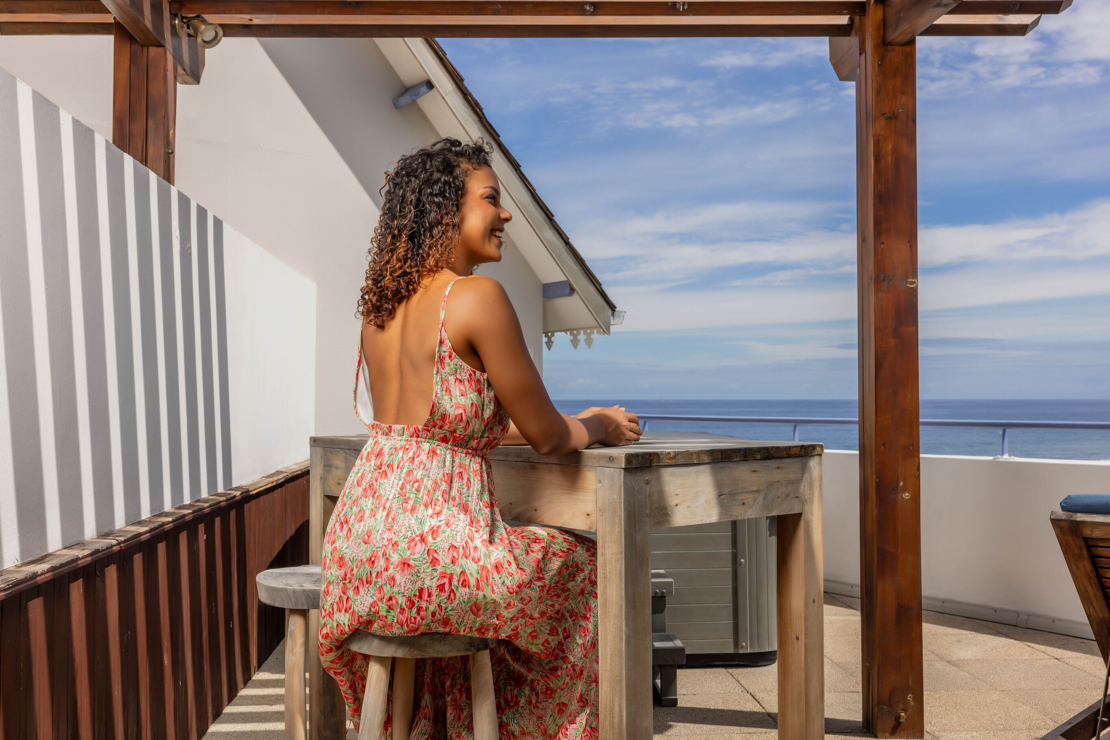 A woman sitting at a wooden table on a balcony overlooking the sea.