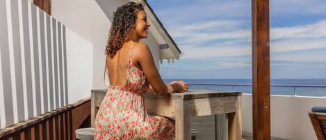A woman sitting at a wooden table on a balcony overlooking the sea.