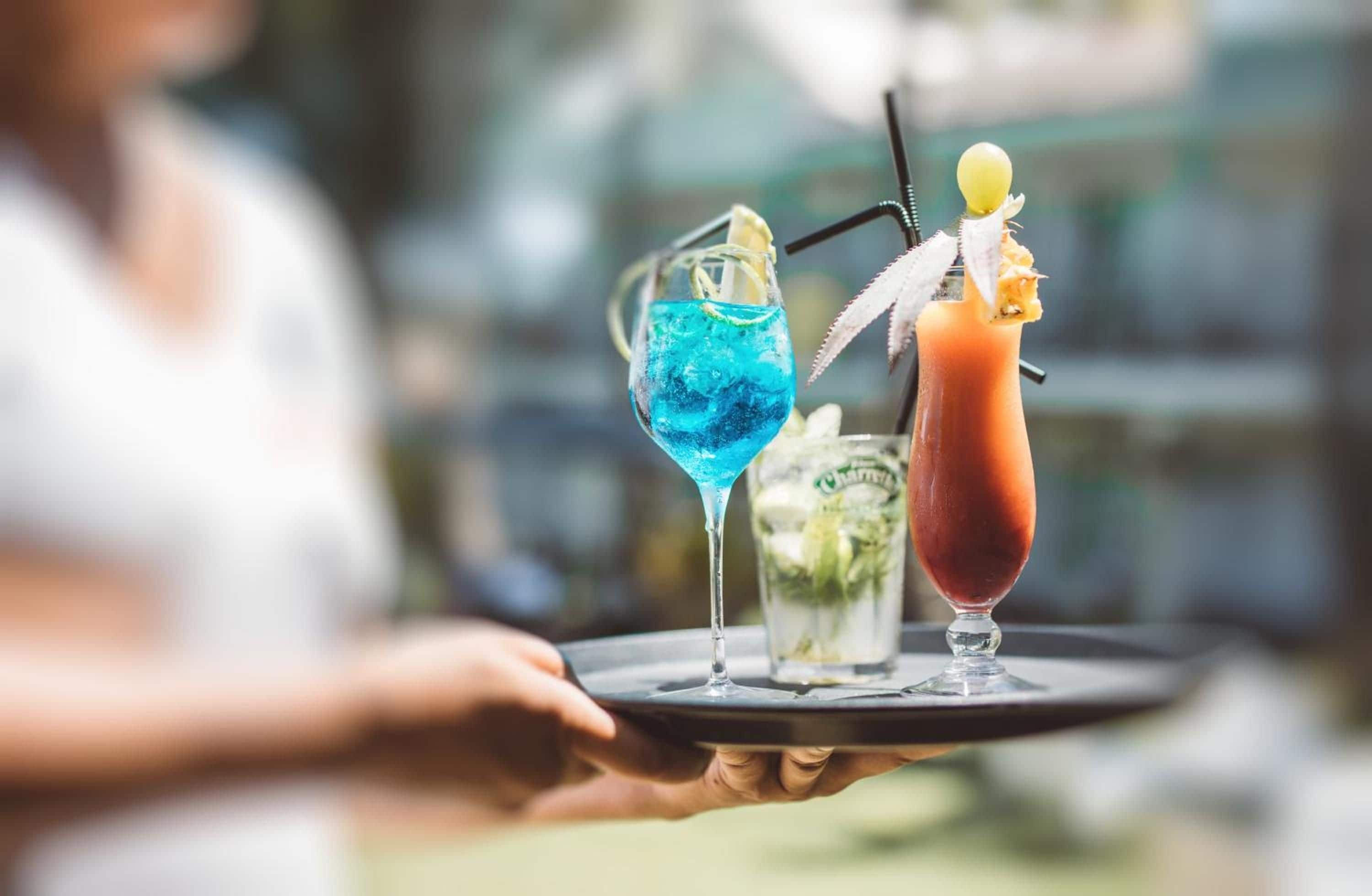 A person holding a tray with two drinks, one blue and one red, each with a straw and garnish.