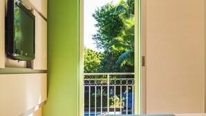 A bedroom with a bed, a television on the wall, and a view of a balcony with greenery. - Hôtel Exsel Alamanda (Saint-Paul)