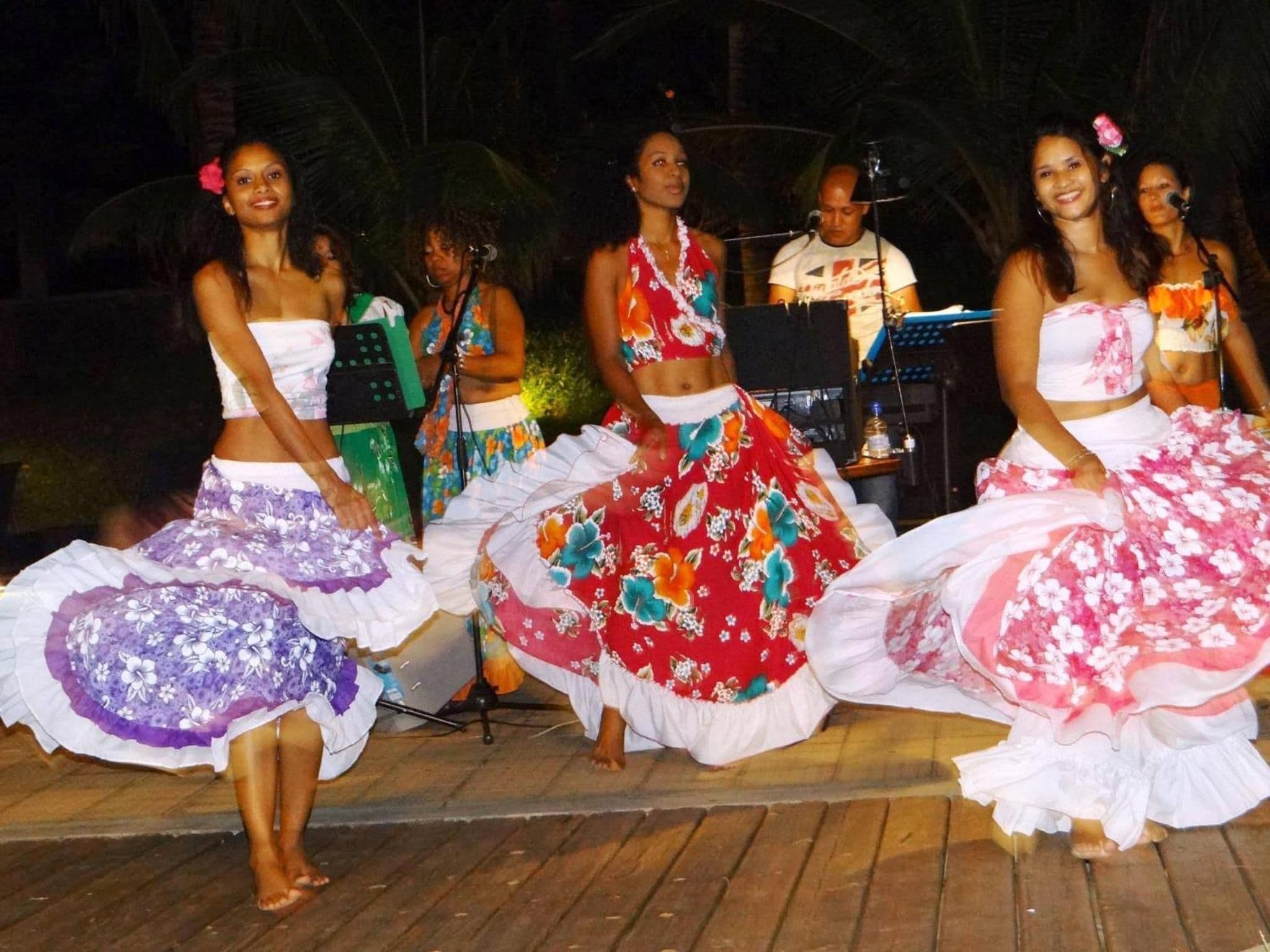 A group of people performing a dance on a wooden stage with a tropical backdrop.