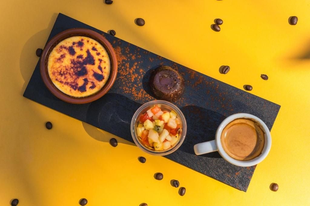 Een dessertplateau met een gekaramelliseerd gerecht, een chocoladedessert en een fruitsalade, vergezeld van een kop koffie op een gele ondergrond.