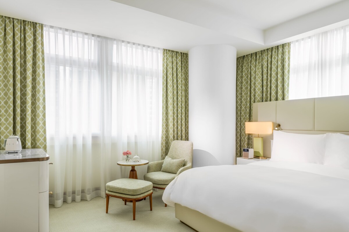 Une chambre d’hôtel avec un lit, une chaise, une petite table avec une lampe et un bureau avec une bouilloire.