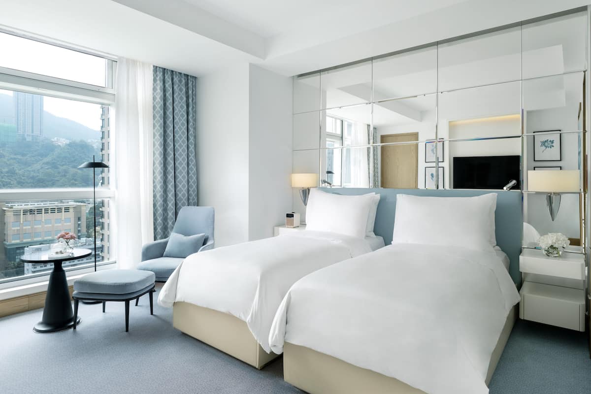 A hotel room with two beds, a chair, a small table, and a large window offering a city view.