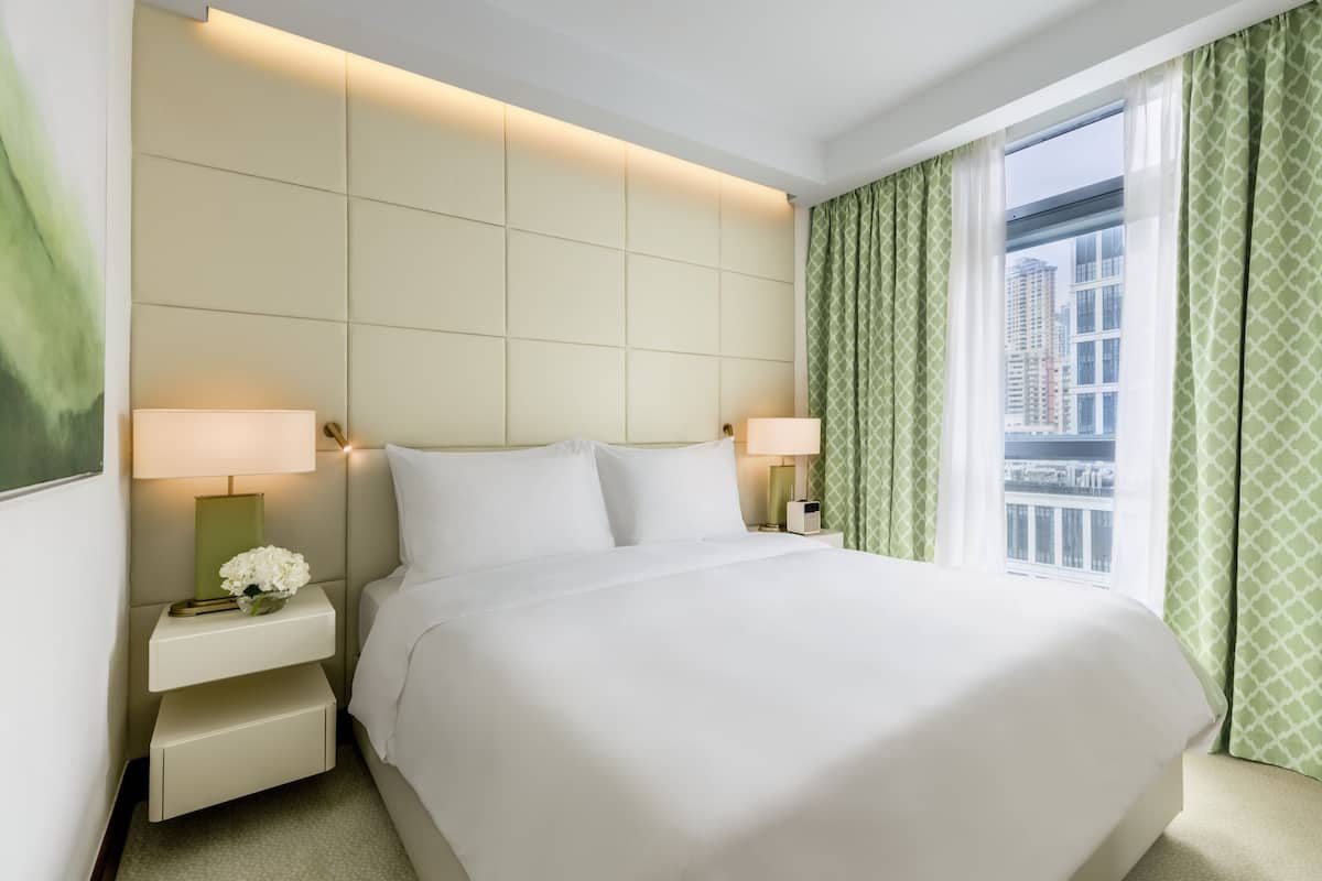A hotel room with a large bed, two bedside tables with lamps, a small table with a vase of flowers, and a window with green curtains.