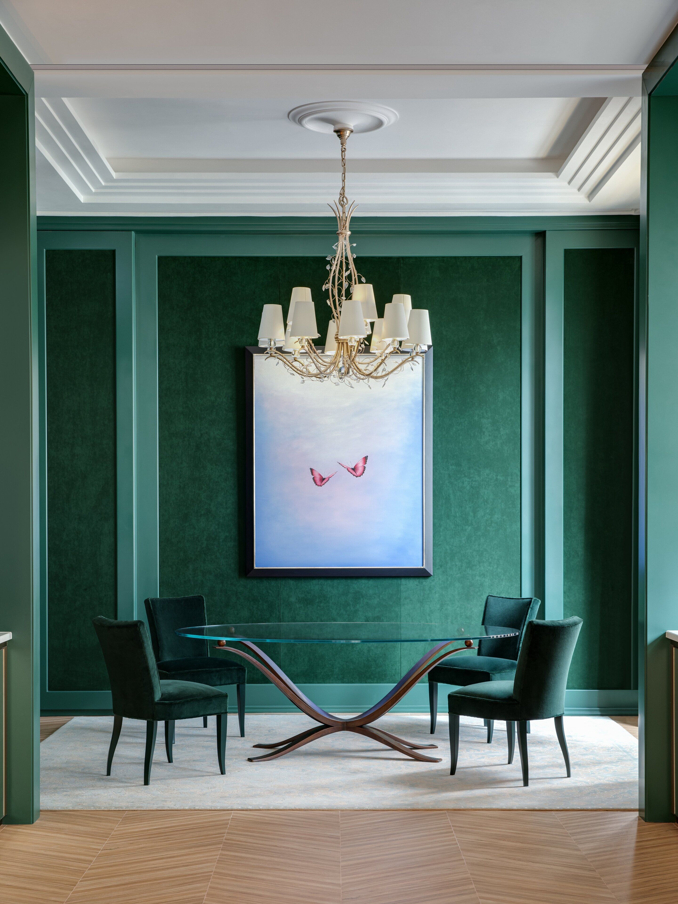 A room with a chandelier, two chairs, a glass table, and a framed painting of birds.