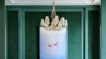 A room with a chandelier, two chairs, a glass table, and a framed painting of birds.