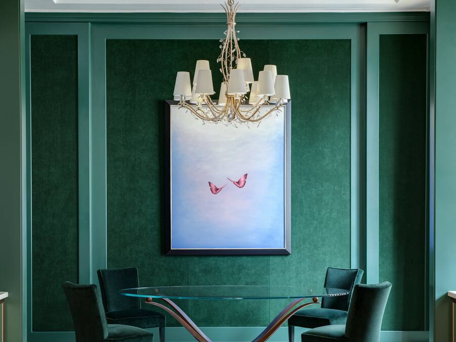 A room with a chandelier, two chairs, a glass table, and a framed painting of birds.