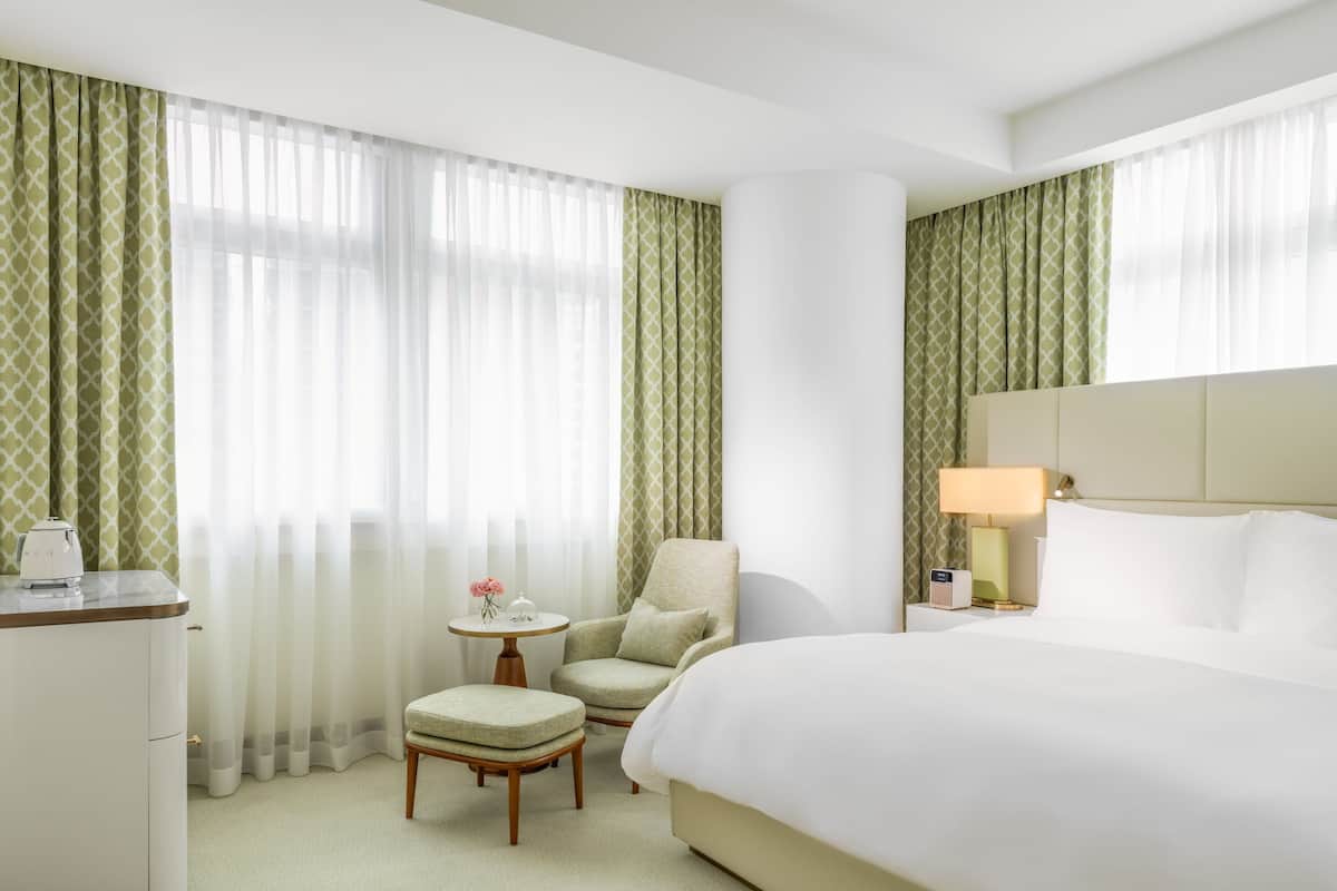 A hotel room with a bed, a chair, a small table with a lamp, and a desk with a kettle.
