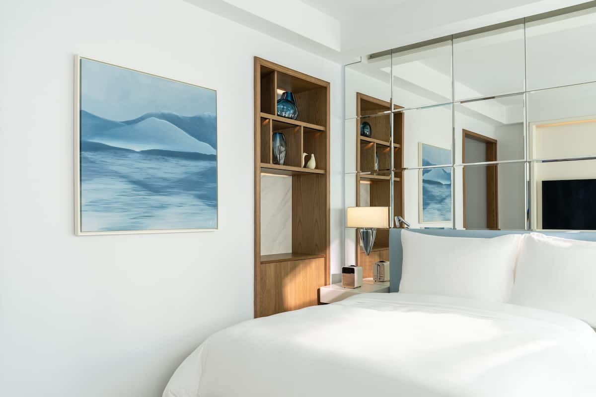 A modern hotel room with a large bed, a wooden shelf with decorative items, a wall-mounted lamp, and a framed picture of a mountain landscape.
