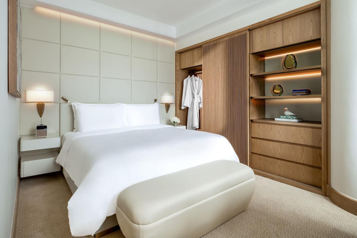 A hotel room with a large bed, bedside lamps, a wooden wardrobe, and a small table with a lamp.