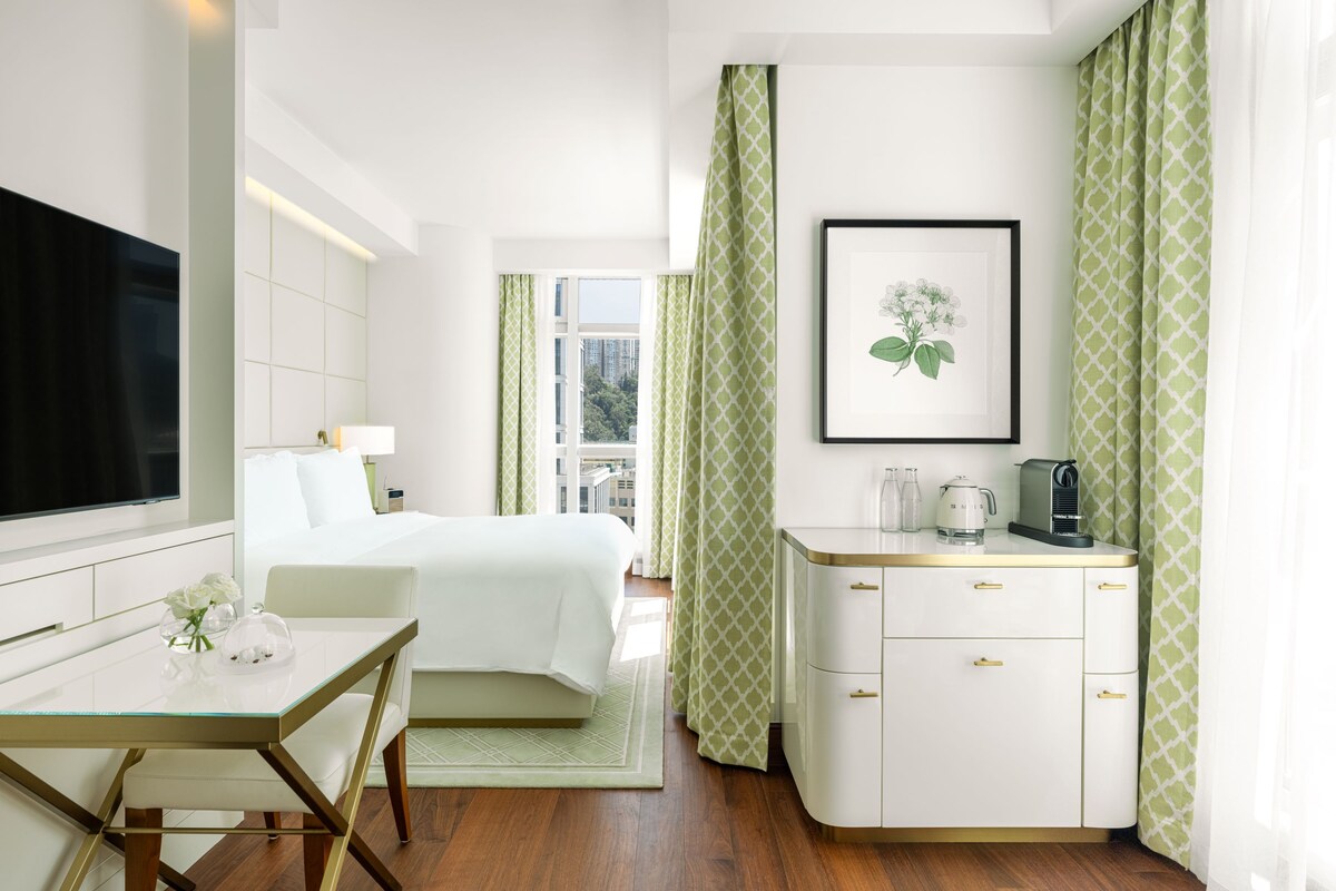 Une chambre d’hôtel avec un lit, un bureau, une télévision, un tableau encadré, une machine à café et une fenêtre avec des rideaux.