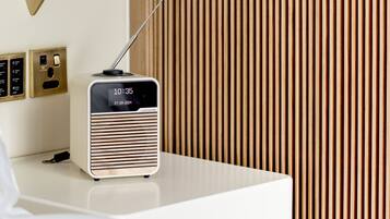 A modern hotel room with a white bedside table, a gold lamp, a radio, and a wooden slat wall.