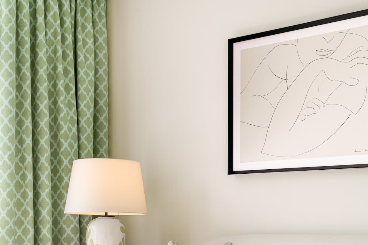 A modern living room with a white sofa, green patterned curtains, a white lamp, and a framed abstract artwork.