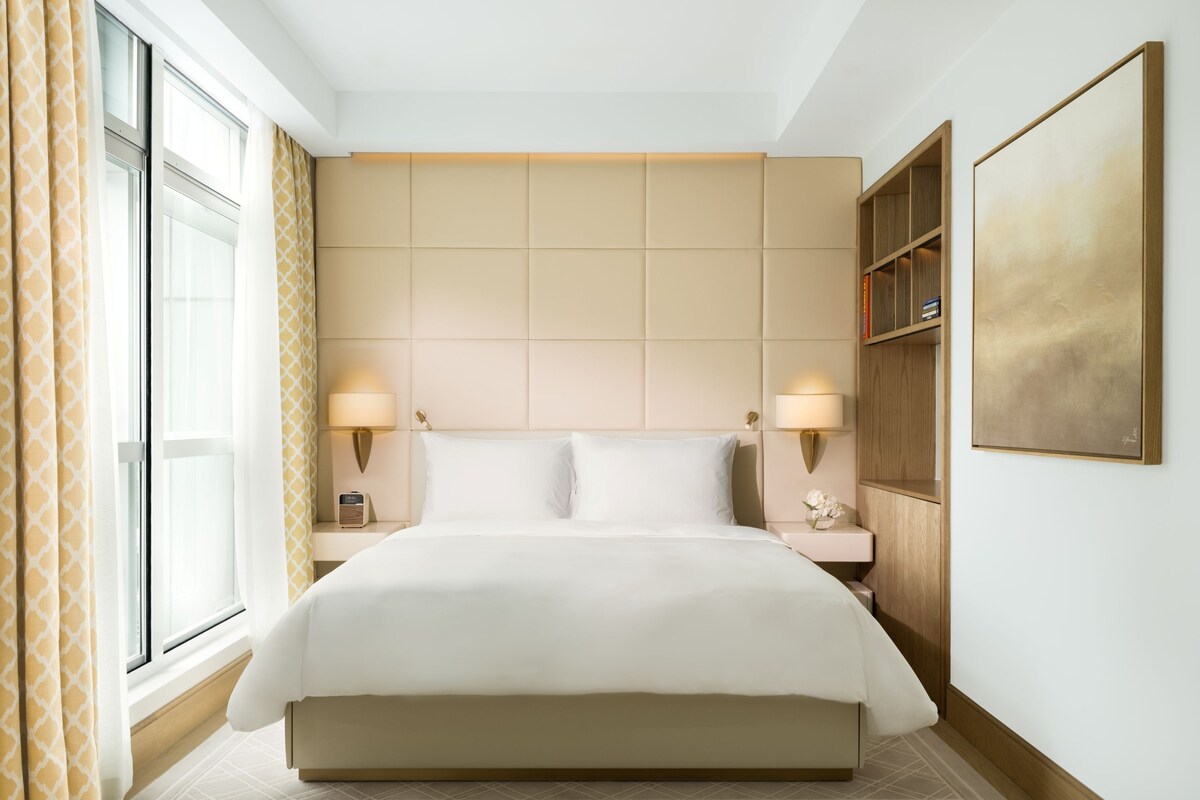 Une chambre d’hôtel avec un grand lit, deux lampes de chevet, une bibliothèque et une grande fenêtre avec des rideaux.