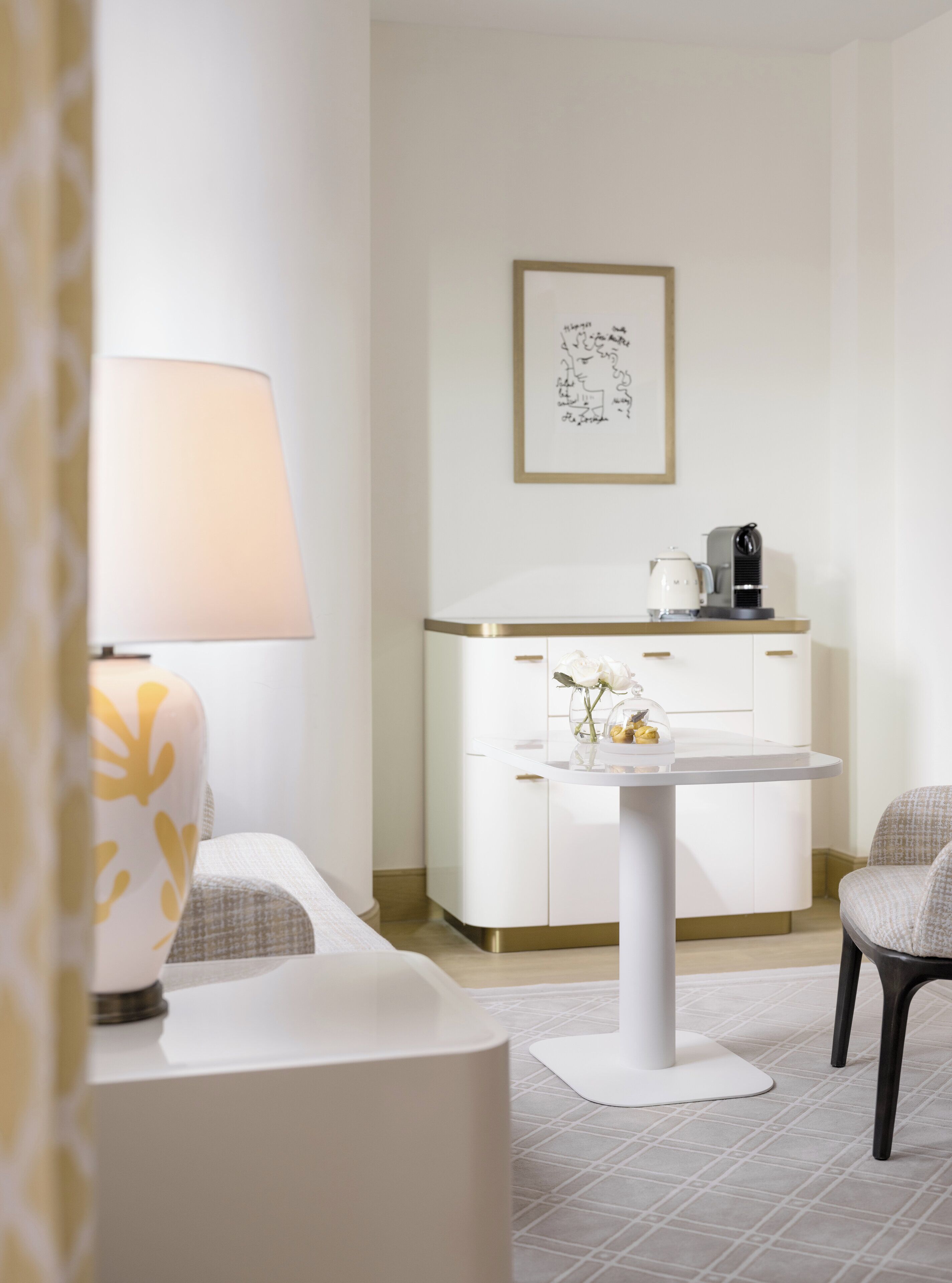 A modern, minimalist room with a white desk, a lamp, a framed artwork, and a coffee machine.