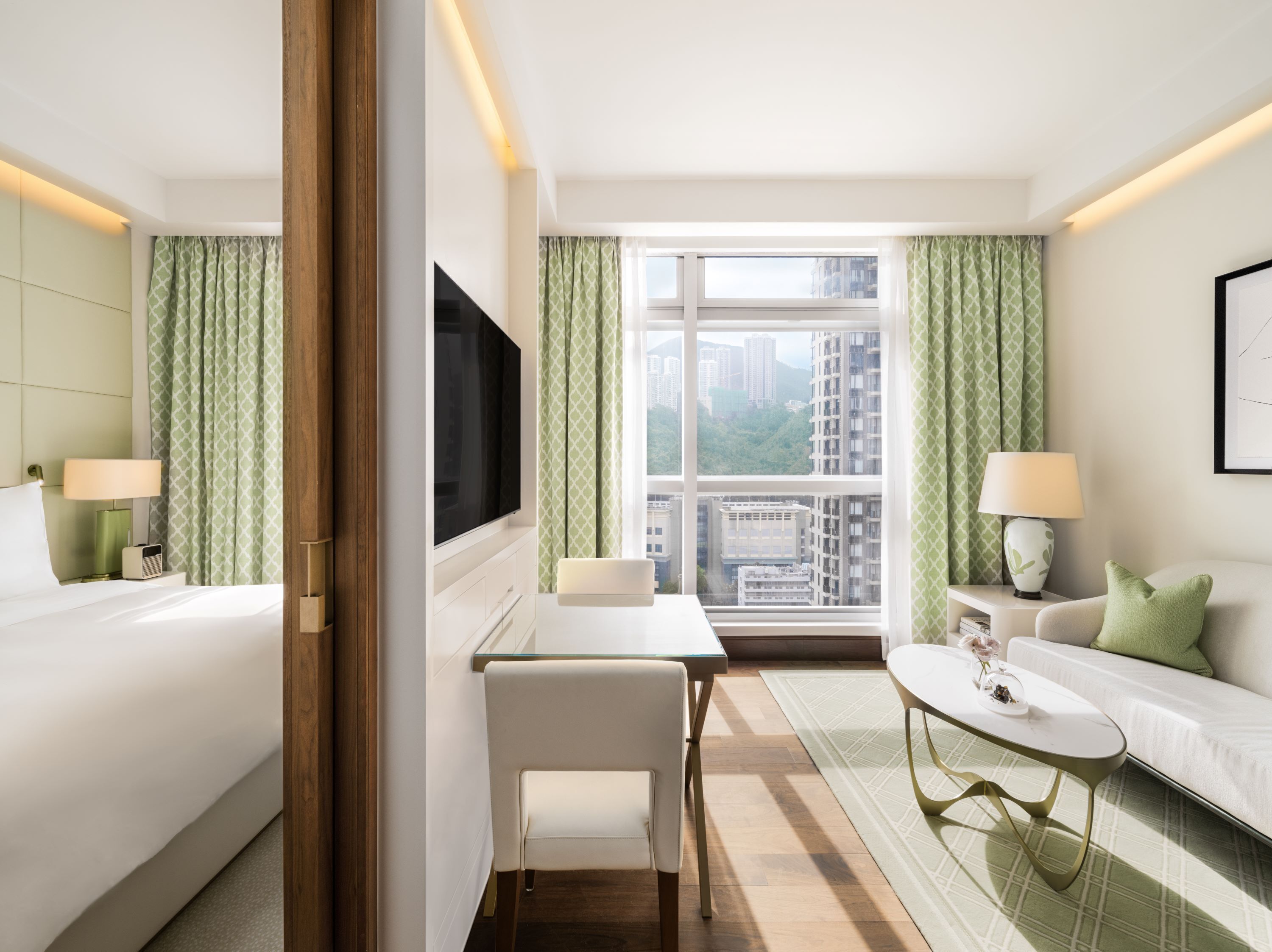 A hotel room with a bed, a desk, a chair, a sofa, a TV, and a window with a city view.