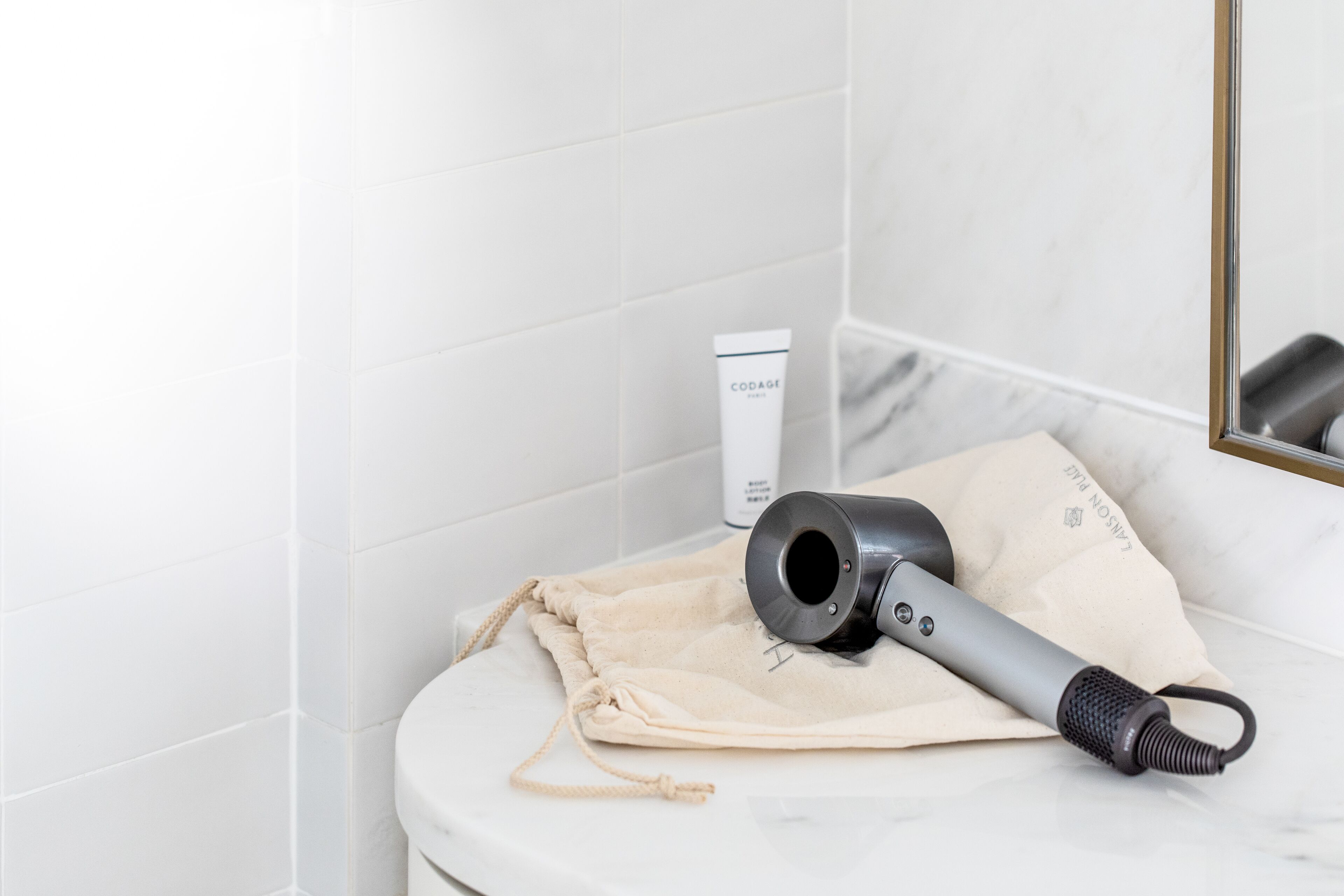 A bathroom with white tiles, a hairdryer, a beige towel, and a bottle of hair product.