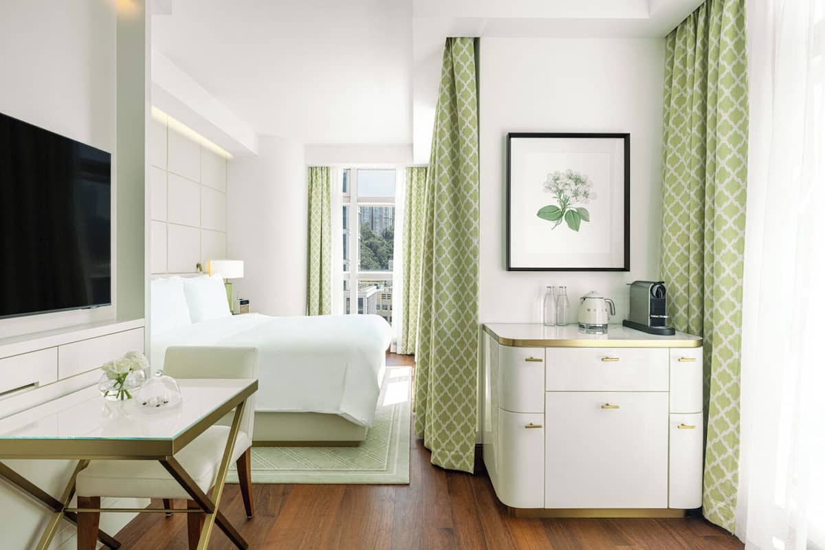 A hotel room with a bed, a desk with a chair, a TV, a framed picture on the wall, and a window with curtains.