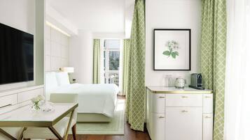 Quarto de hotel com cama, escrivaninha com cadeira, televisão, quadro na parede e janela com cortinas.
