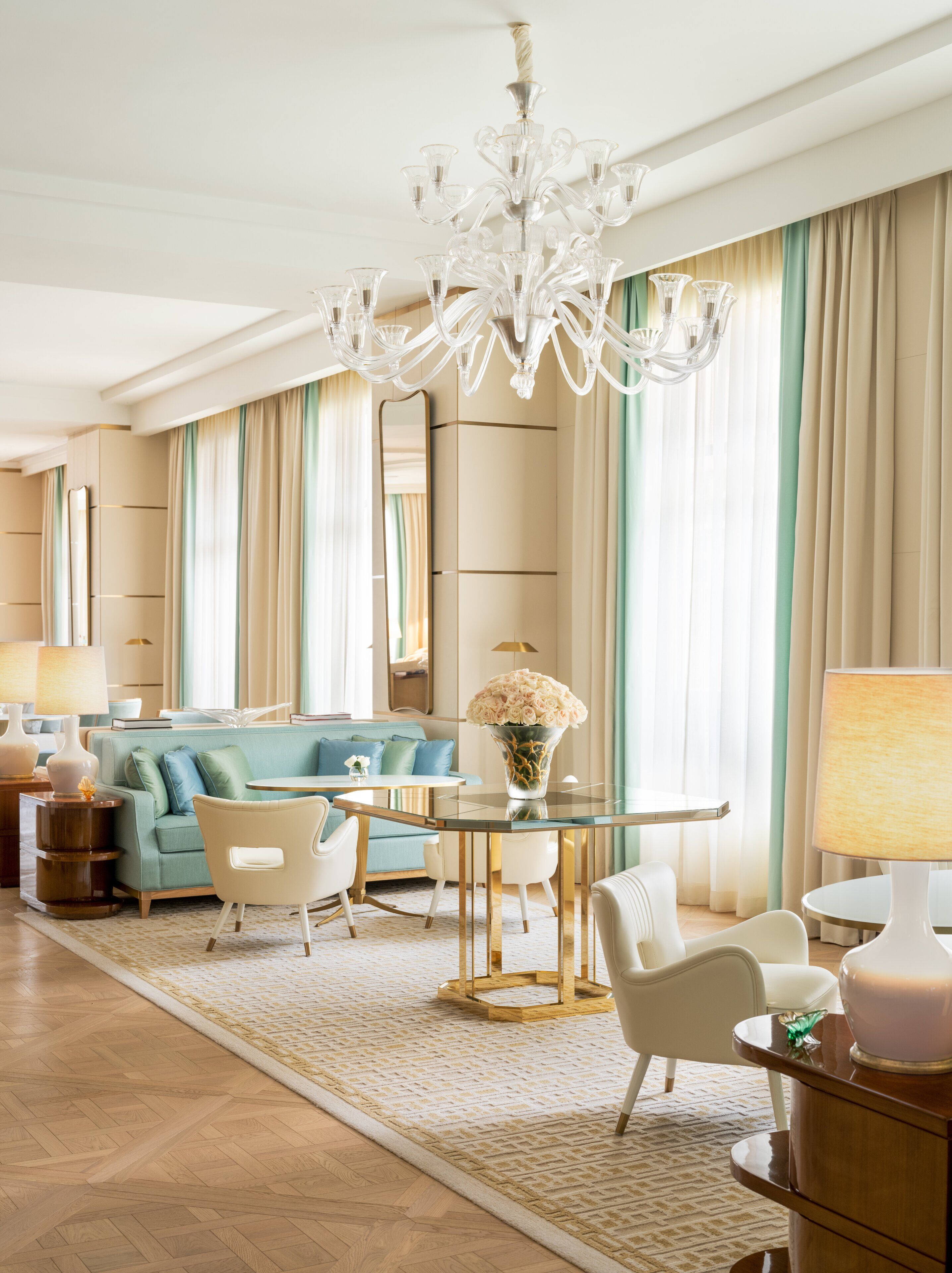 A spacious living room with a chandelier, a blue sofa, a round glass table, and a large mirror.