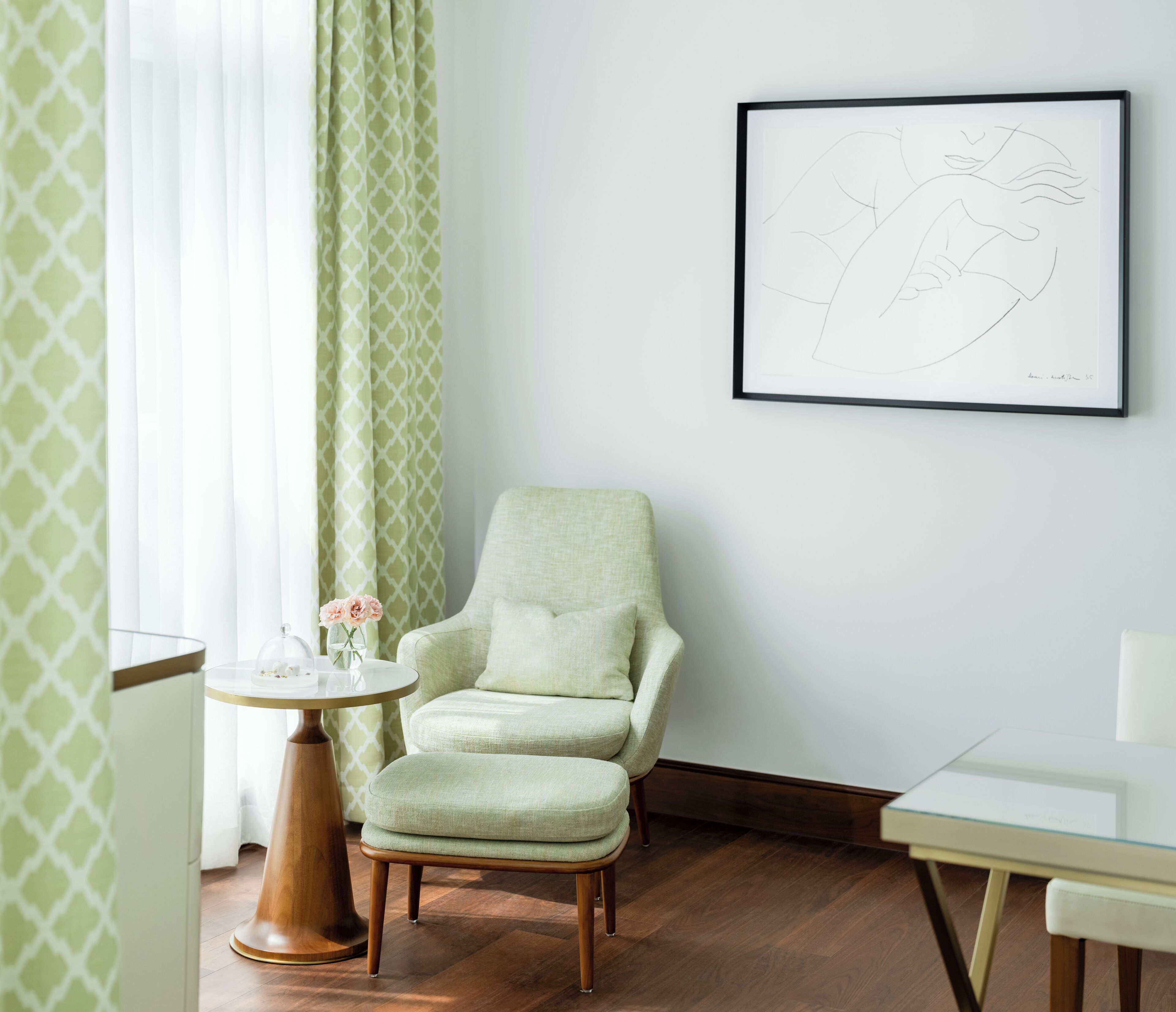 A room with a green armchair, a small round table with a vase of flowers, and a framed artwork on the wall.