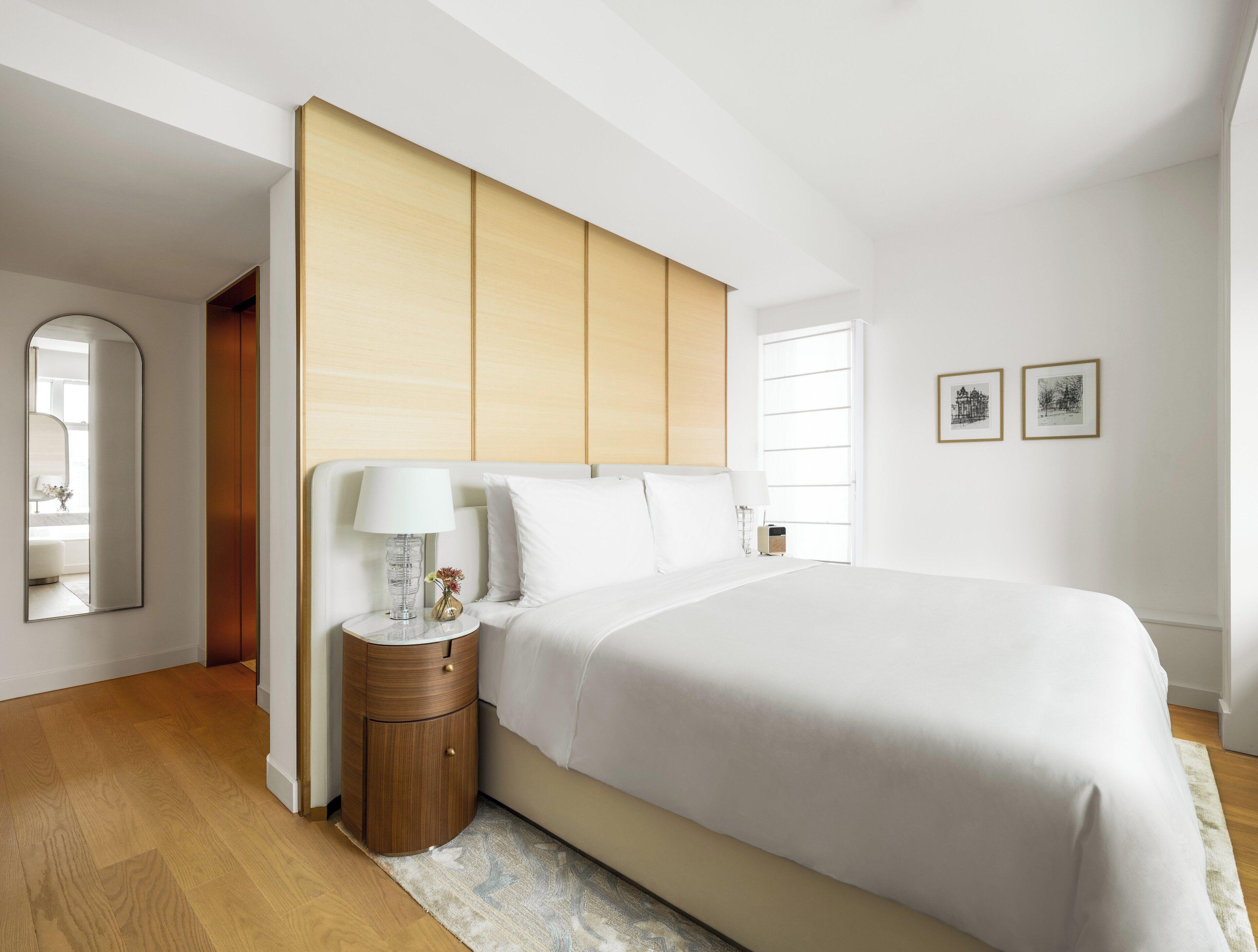 Un dormitorio moderno con una cama grande, una mesita de noche con lámpara, un espejo y dos cuadros en la pared.