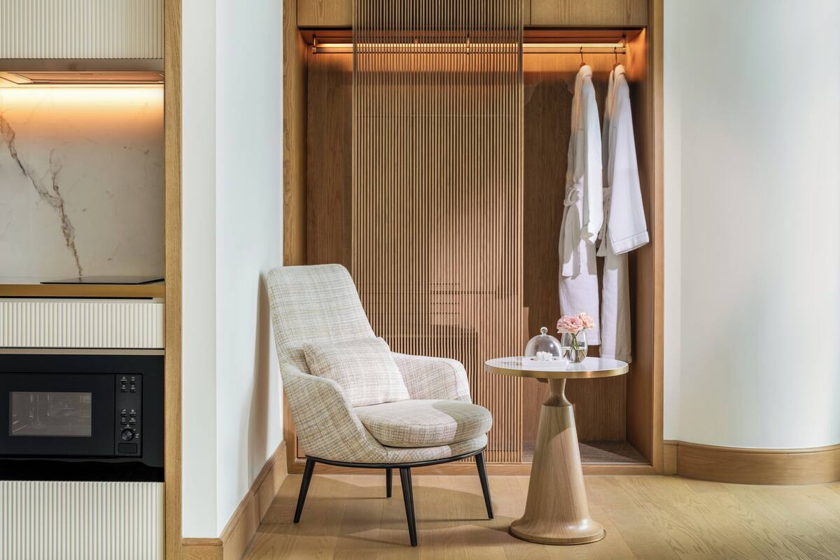 A modern bathroom with a wooden vanity, a white chair, a small round table with a vase of flowers, and a towel rack with towels.