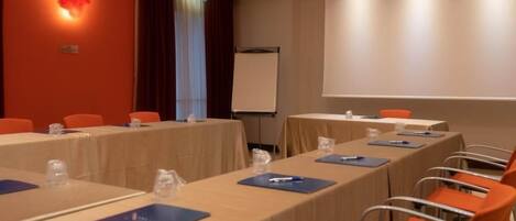 Un salón de conferencias con largas mesas, sillas anaranjadas y una pantalla para proyector.