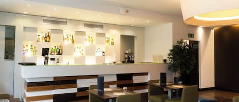 Un espace bar moderne avec un comptoir où sont exposées différentes bouteilles, un coin salon avec des tables et des chaises, et une grande plante en pot.