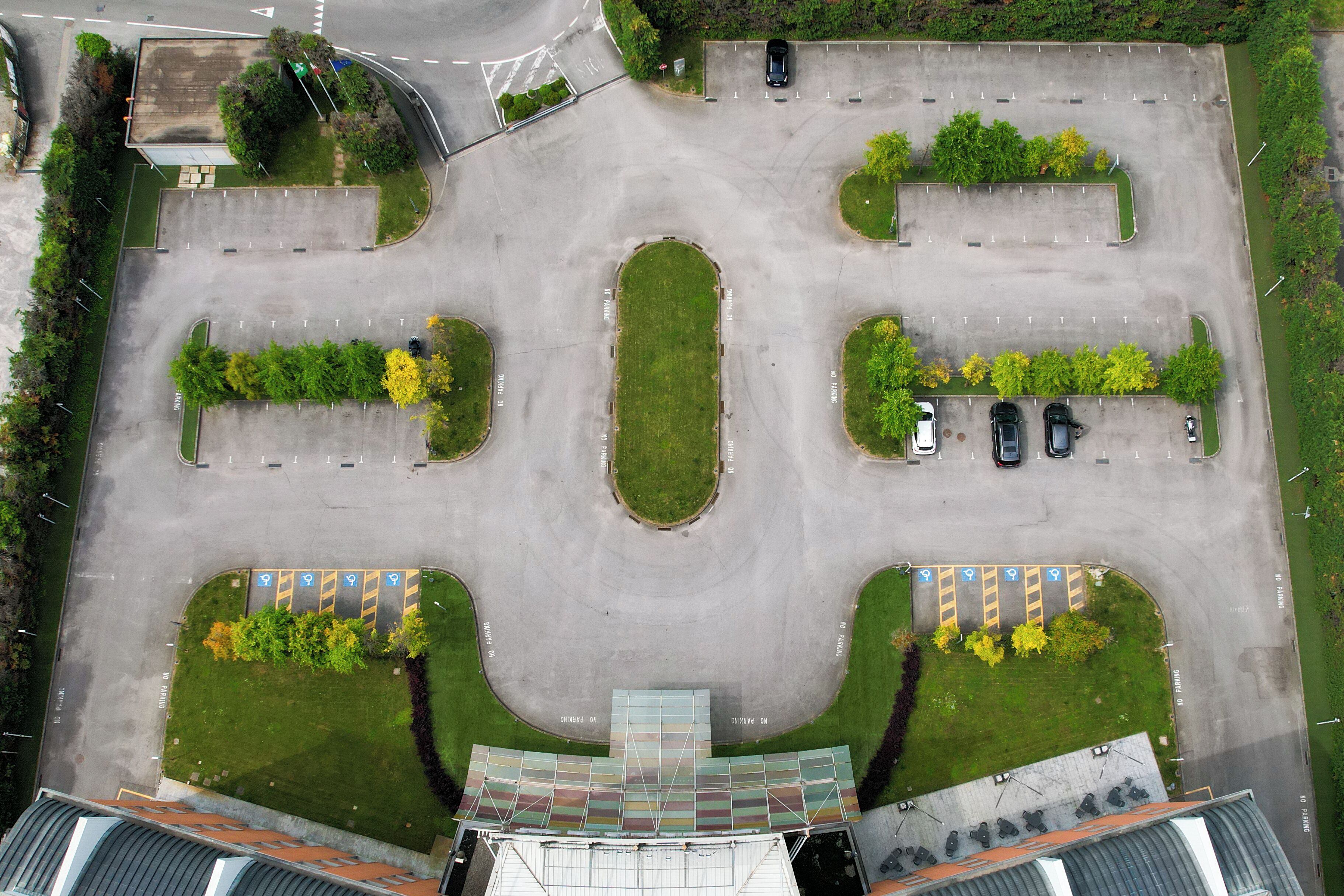 An aerial view of a parking lot with multiple parking spaces, green landscaping, and a building with a curved roof in the foreground.