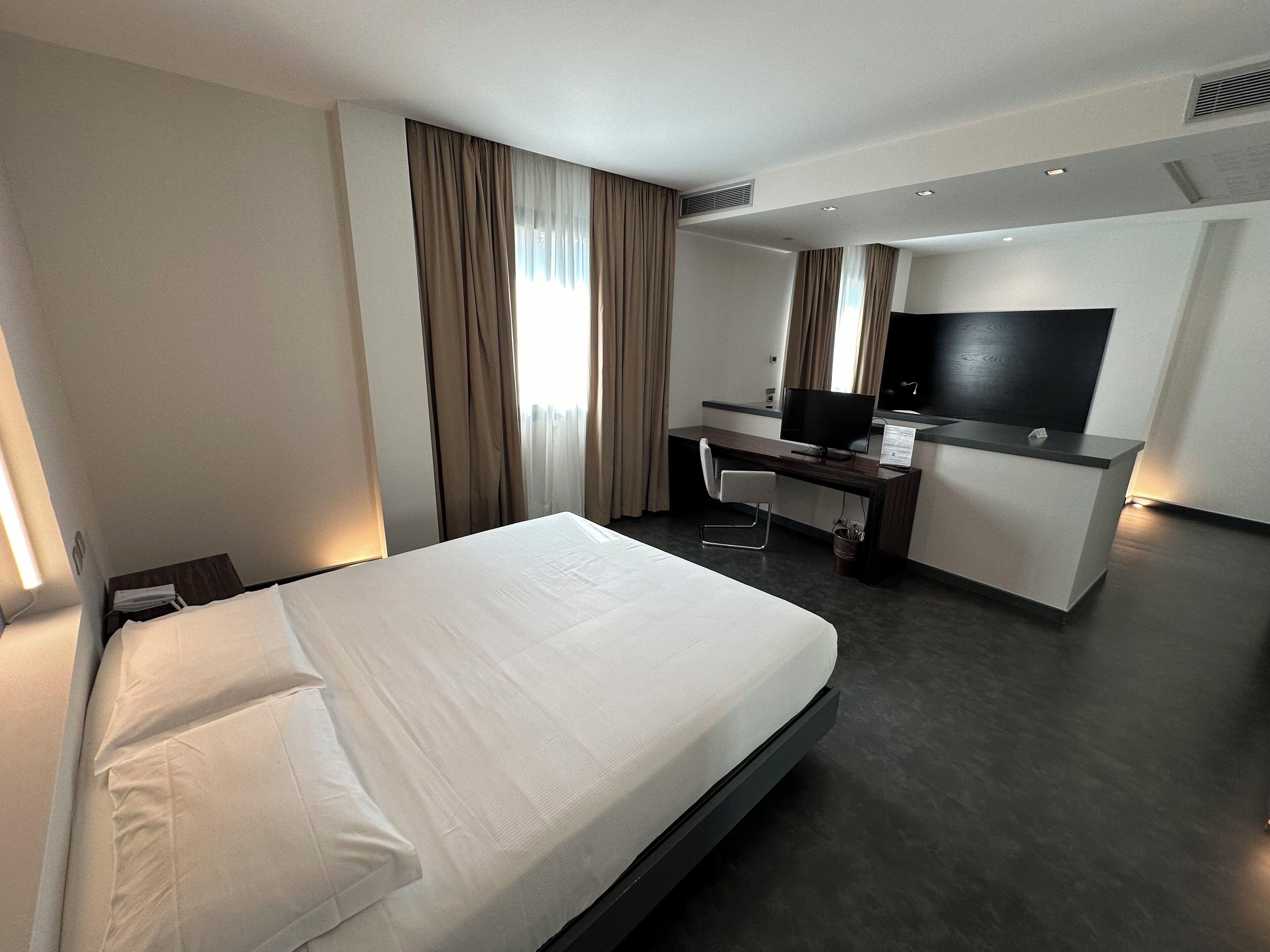 A hotel room with a bed, a desk with a computer, a TV, and a window with curtains.