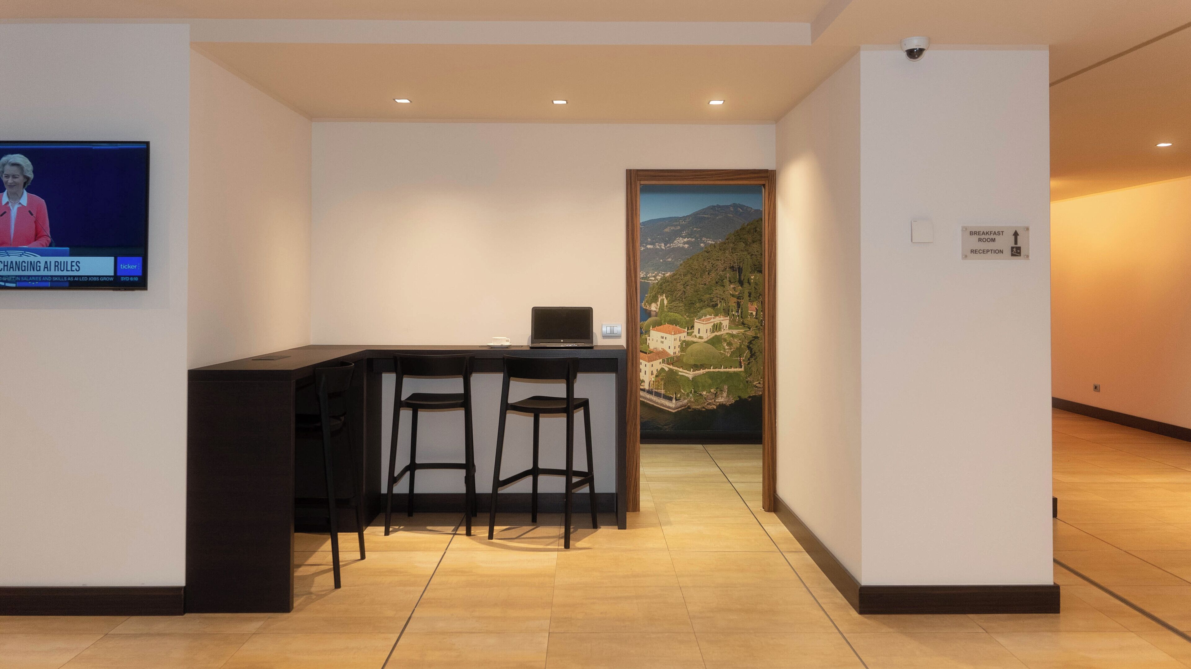Un área de recepción con un mostrador, dos sillones, un televisor mostrando un ticker de noticias y un cuadro con un paisaje.
