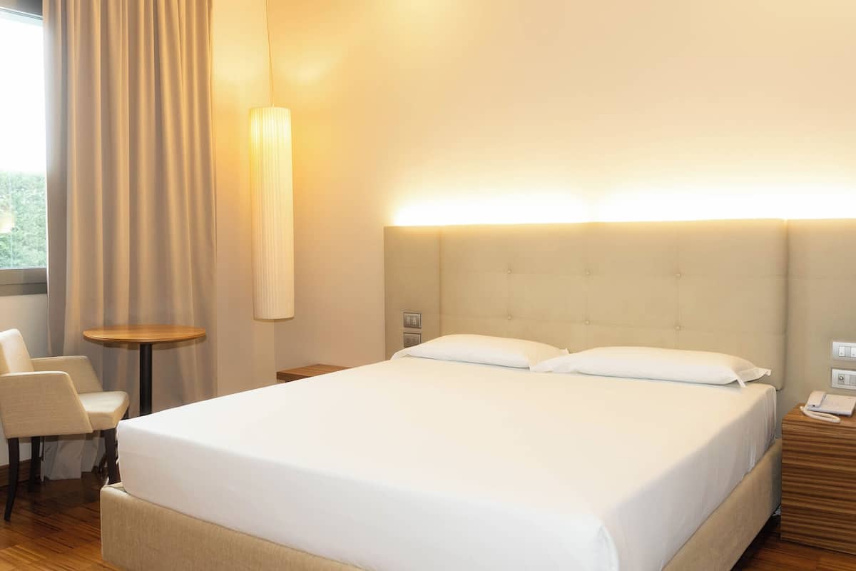 A hotel room with a large bed, a chair, a small table, and a bedside table with a lamp.
