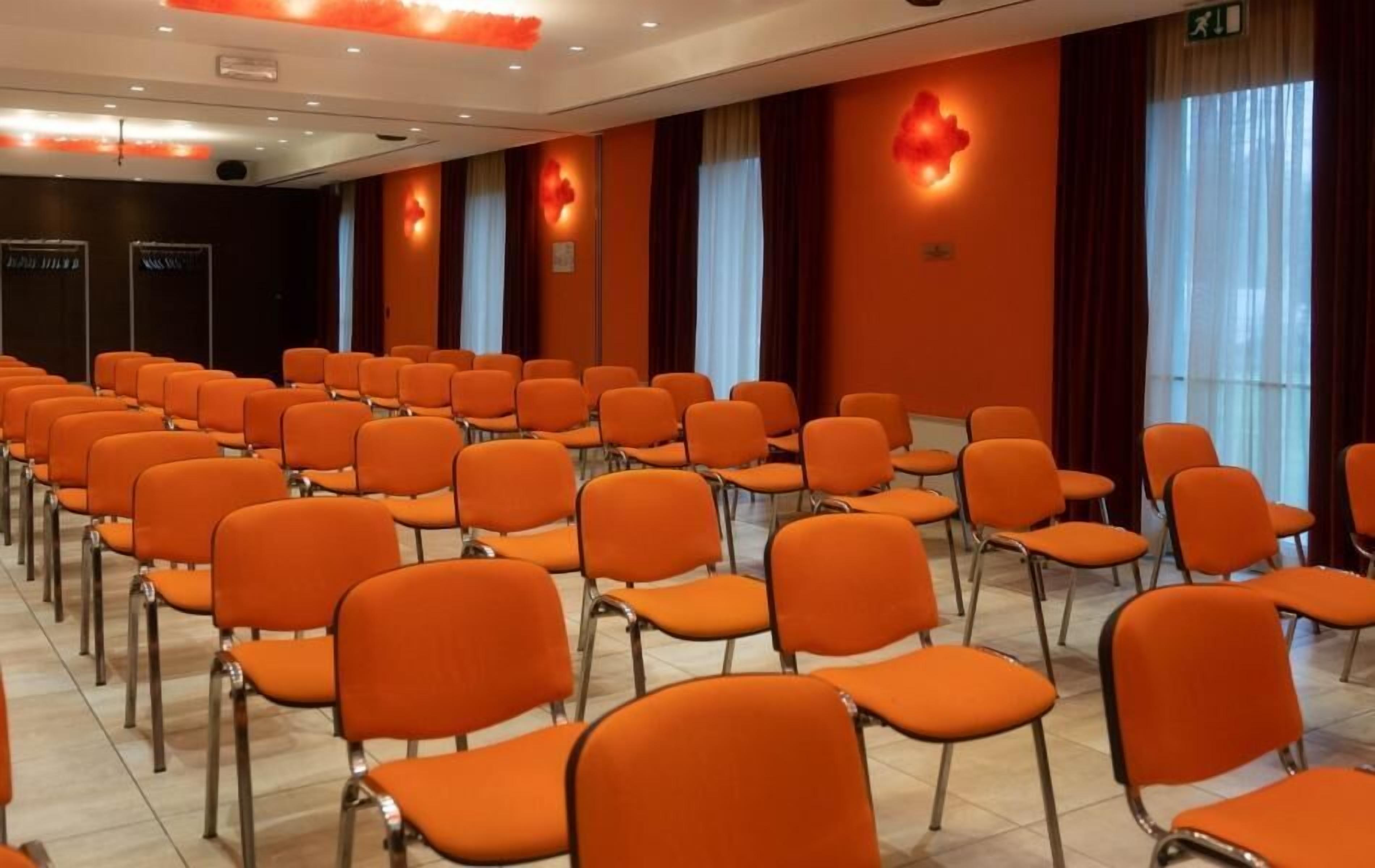 Un salón de conferencias con filas de sillas naranjas, un escenario y un amplio ventanal con cortinas.