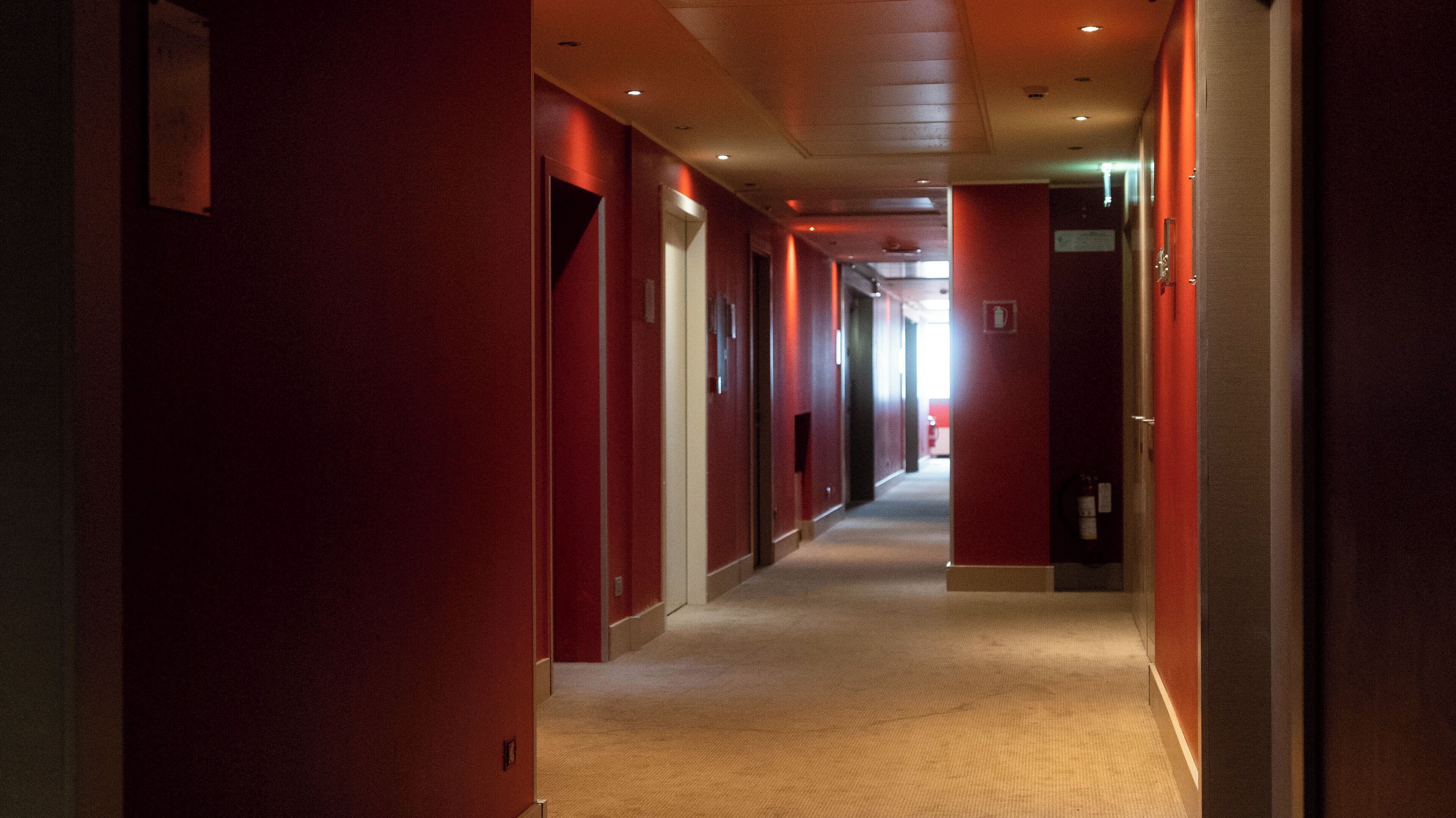 A long, narrow corridor with red walls and a carpeted floor, leading to a bright exit at the end.