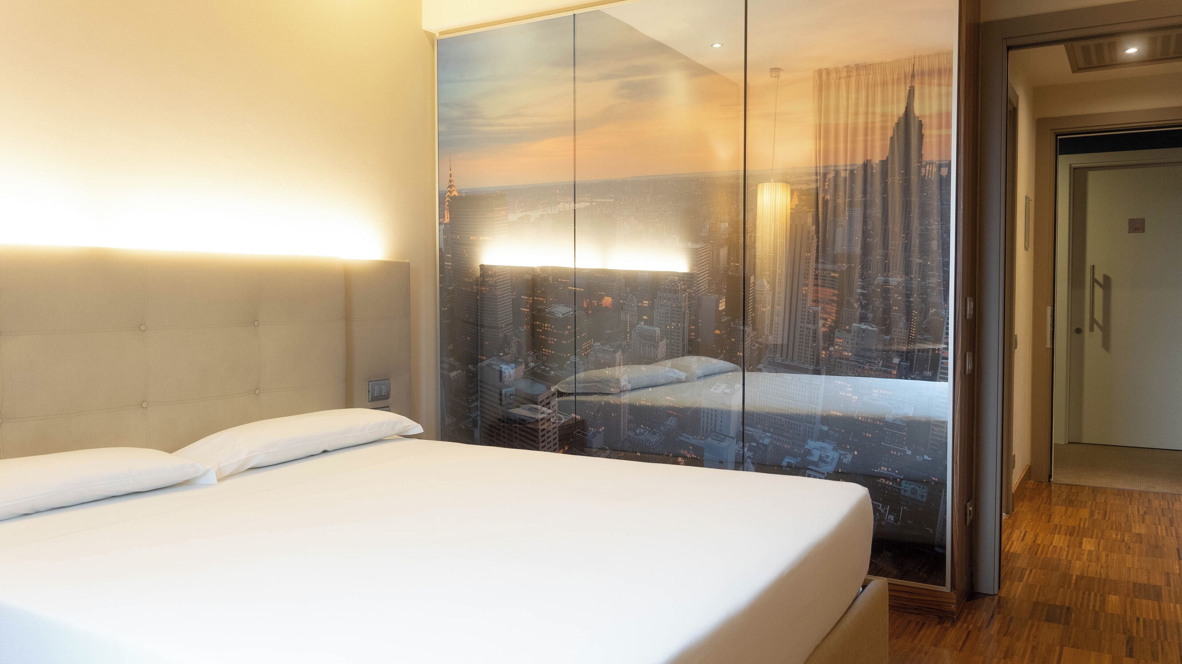 A hotel room with a large bed, a glass partition wall, and a view of a cityscape.
