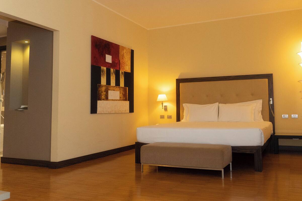 A hotel room with a large bed, a bedside table, a nightstand, a wall-mounted lamp, and a painting.