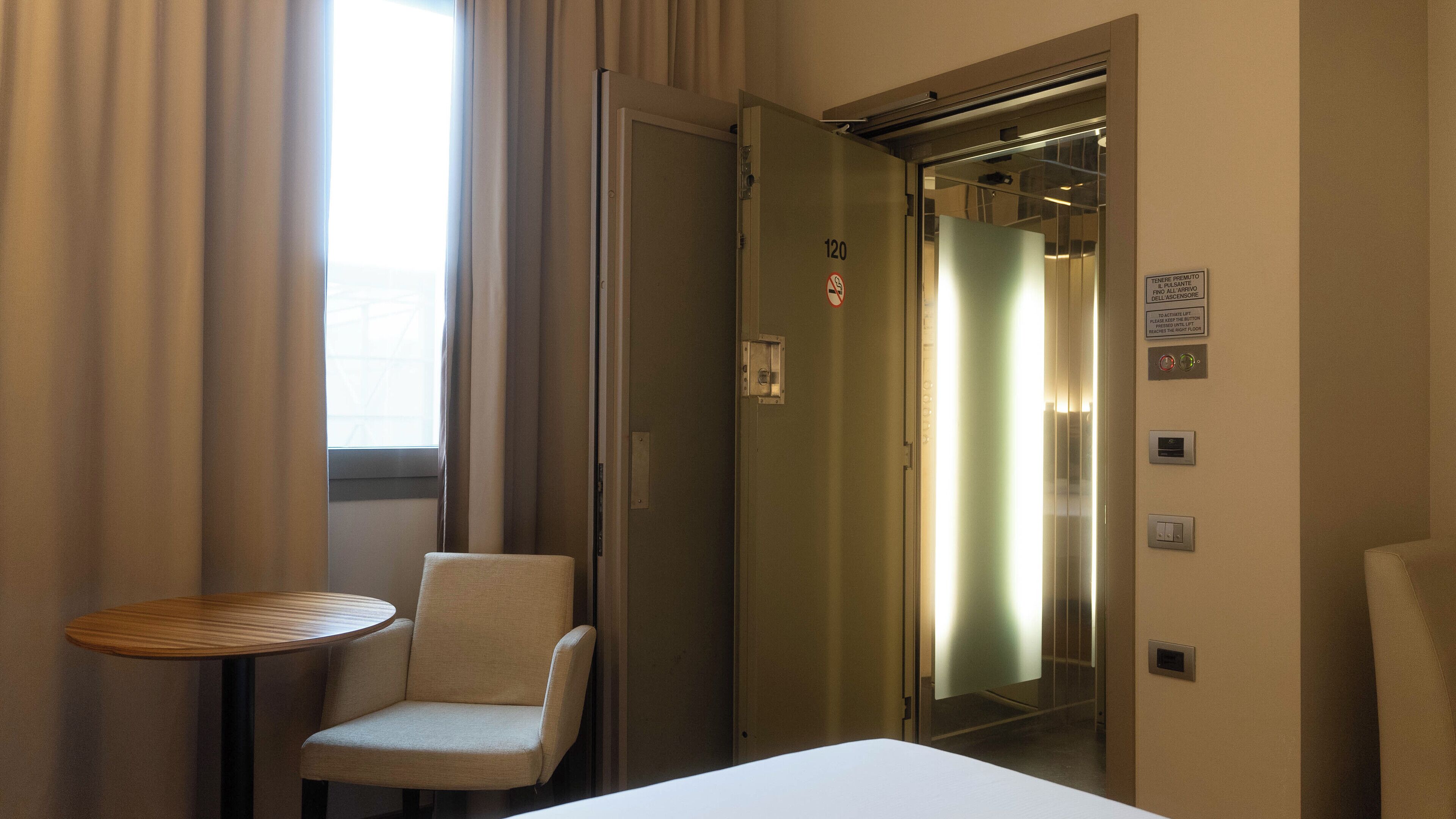 A hotel room with a bed, a chair, a small table, and an elevator.