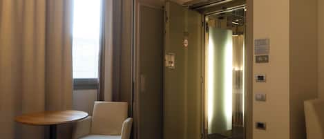 A hotel room with a bed, a chair, a small table, and an elevator.