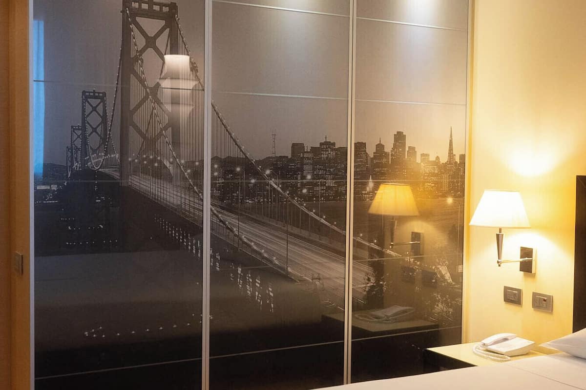 A hotel room with a large bed, a cityscape view through a sliding door, a wall-mounted lamp, and a wooden floor.