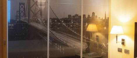 A hotel room with a large bed, a cityscape view through a sliding door, a wall-mounted lamp, and a wooden floor.