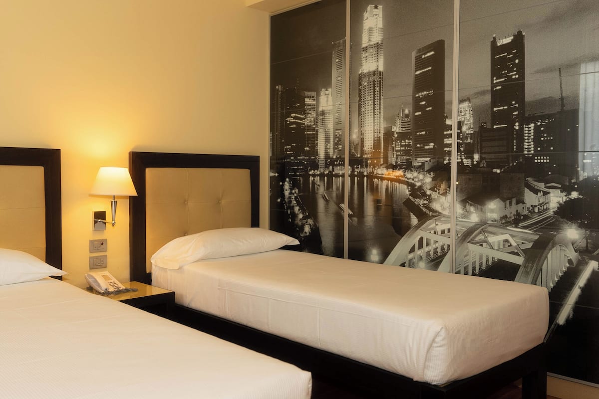 A hotel room with two single beds, a nightstand with a lamp, a phone, and a large cityscape artwork on the wall.