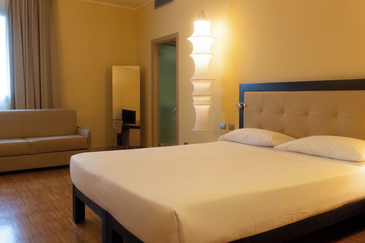 A hotel room with a large bed, a chair, a desk, and a wall-mounted lamp.