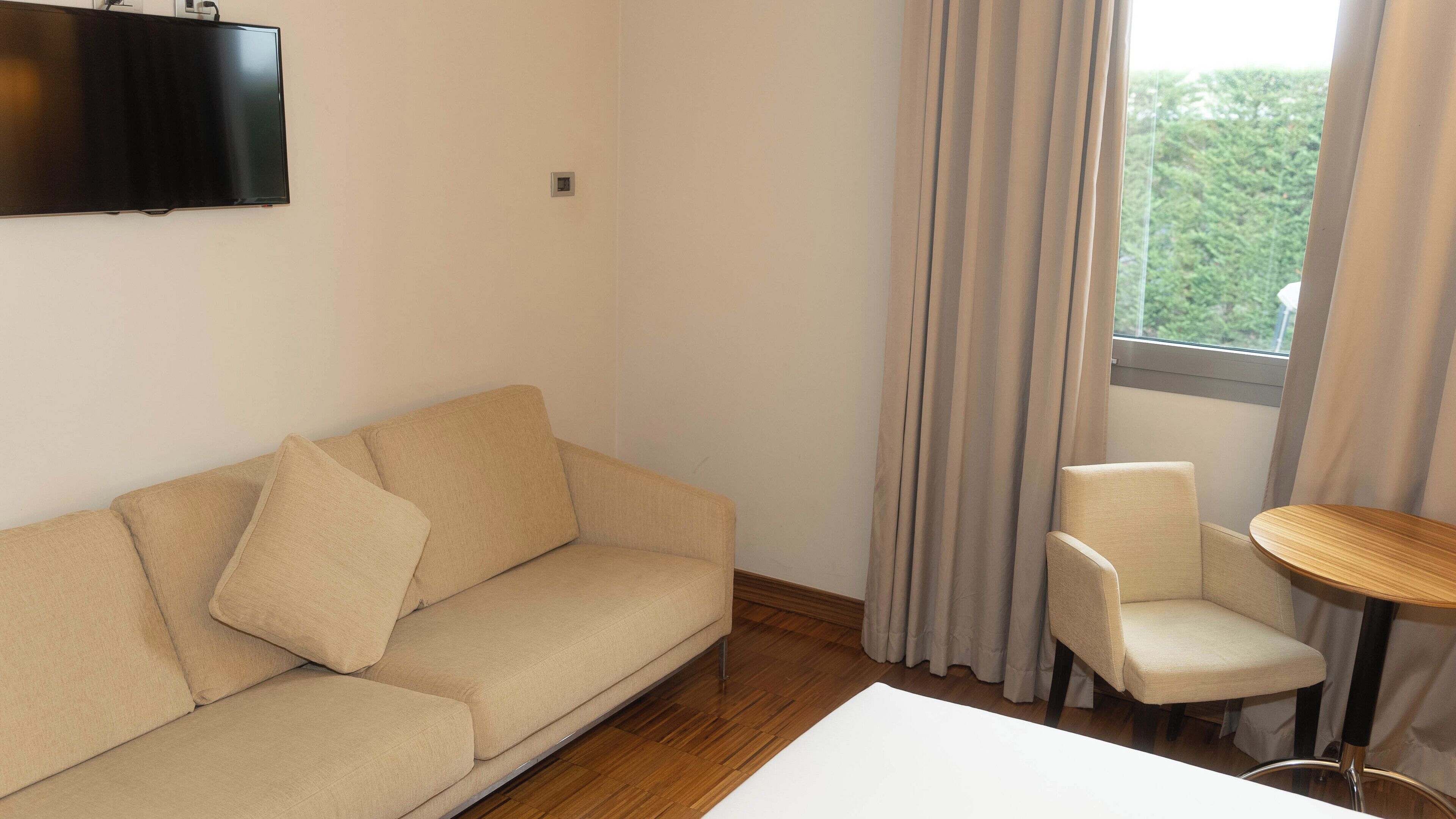Ein Zimmer mit einem beigefarbenen Sofa, einem kleinen runden Tisch und einem an der Wand befestigten Fernseher.